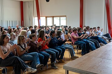 Schülerinnen und Schüler klatschen bei einer Veranstaltung in einem hellen Raum mit Holzboden und roten Säulen.